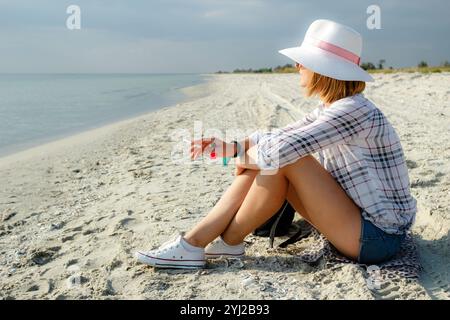 Eine junge Frau hält draußen eine Zigarette in der Hand. Porträt einer jungen Frau mit weißem Hut, die am Meer sitzt und eine Zigarette raucht. Smoki Stockfoto