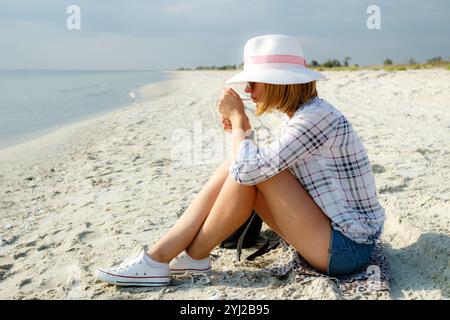 Porträt einer jungen Frau mit weißem Hut, die am Meer sitzt und eine Zigarette anzündet. Rauchen Eine junge Frau raucht draußen eine Zigarette. Konz Stockfoto