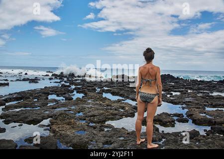 Eine Frau im Bikini steht an einer felsigen Küste und blickt auf das Meer, während die Wellen gegen die Felsen krachen. Stockfoto