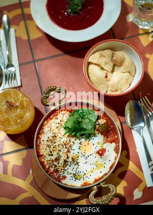 Von oben aus bietet sich eine lebhafte Mahlzeit aus dem Nahen Osten mit Shakshuka, Rote-Bete-Suppe, Pitabrot und einem Orangengetränk auf einem gemusterten Tisch. Stockfoto