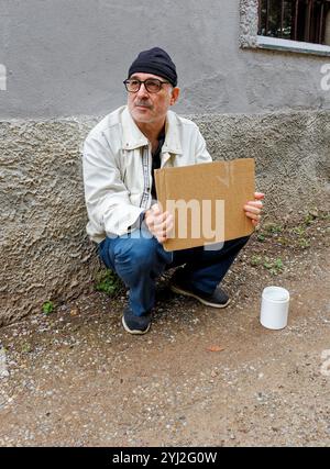 Älterer Mann mit Bart, Baskenmütze und Brille, sitzt auf dem Bürgersteig und hält ein Pappschild neben einem weißen Becher, um Geld zu sammeln, Italien Stockfoto