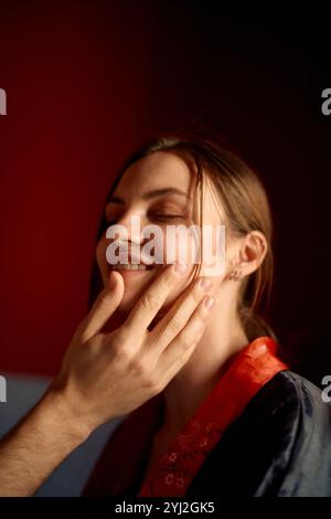 Lächelnde Frau mit geschlossenen Augen, die ihr Gesicht in einem sanft beleuchteten Raum sanft berührt. Stockfoto