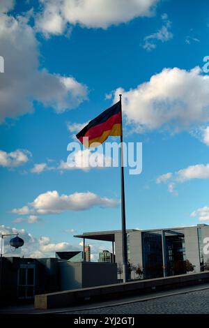 Eine deutsche Flagge flattert im Wind vor einem Hintergrund von blauem Himmel und verstreuten Wolken, mit modernen Gebäuden in der Ferne, Berlin, Deutschland Stockfoto