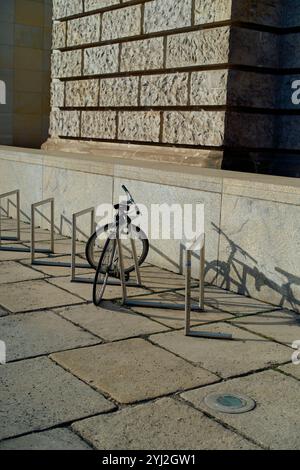 Ein einzelnes Fahrrad, das an einem Fahrradträger aus Metall befestigt ist, wirft an einem sonnigen Tag in der Nähe einer Steinmauer einen langen Schatten Stockfoto