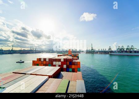 Ein Frachtschiff, beladen mit bunten Containern, fährt an einem klaren Tag mit einem geschäftigen Hafen im Hintergrund, Singapur, ins Meer Stockfoto