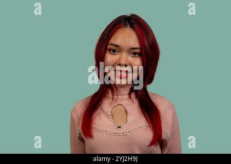Studio-Porträt einer fröhlichen jungen lateinfrau mit gefärbten roten Haaren, die einen modischen rosa Pullover trägt und vor einem leuchtenden türkisfarbenen Hintergrund posiert Stockfoto