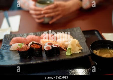 Hände, die eine Tasse mit Sushi-Sortiment im Vordergrund auf einem schwarzen Teller wiegen, begleitet von Ingwer, Wasabi und Miso-Suppe. Stockfoto