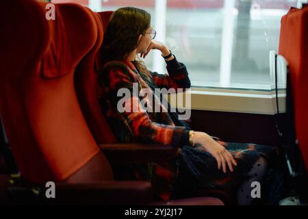 Frau in Brille sitzt bequem auf einem roten Zugsitz und blickt tief in Gedanken aus dem Fenster. Stockfoto