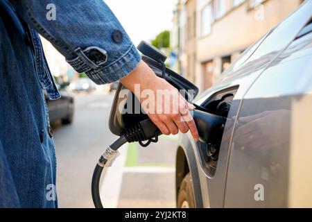 Nahaufnahme einer Person, die an einem sonnigen Tag einen Ladestecker in den Ladeanschluss eines Elektrofahrzeugs einsteckt. Stockfoto