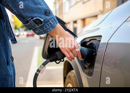 Nahaufnahme einer Person, die an einem sonnigen Tag einen Ladestecker in den Ladeanschluss eines Elektrofahrzeugs einsteckt. Stockfoto