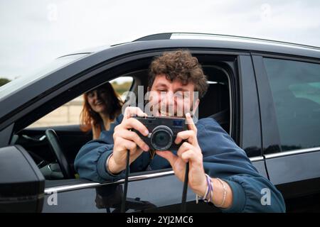Lässiger und fröhlicher bärtiger Mann, der ein Foto mit einer Digitalkamera aus dem Fenster eines Autos mit einer lächelnden Frau im Hintergrund macht. Stockfoto