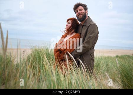 Lächelndes Paar umarmt in hohem Gras mit einem ruhigen Strand im Hintergrund, Belgien Stockfoto