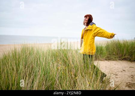 Frau in gelbem Regenmantel genießt die Ruhe an einem Strand mit hohem Gras, Belgien Stockfoto