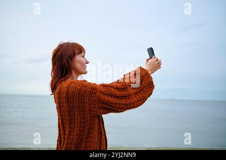 Frau in einem gestrickten orangefarbenen Pullover, die an einem bewölkten Strandtag ein Selfie mit ihrem Handy macht, Belgien Stockfoto