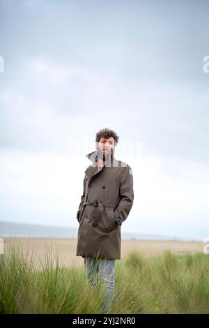 Mann in braunem Mantel auf einem Feld mit hohem Gras und bewölktem Himmel im Hintergrund, Belgien Stockfoto