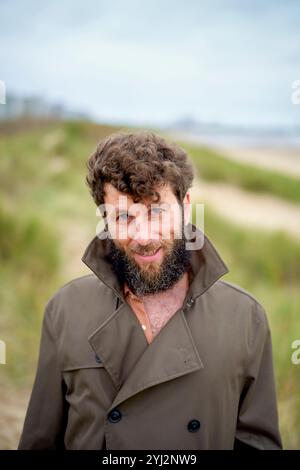 Lächelnder Mann mit lockigen Haaren und Bart, der an einem Strand mit Sanddünen im Hintergrund steht, Belgien Stockfoto