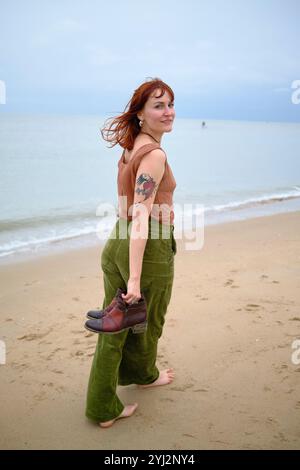 Frau mit rotem Haar und einem Tattoo auf dem Arm geht barfuß an einem Sandstrand, hält Schuhe in einer Hand und schaut über die Schulter, Belgien Stockfoto