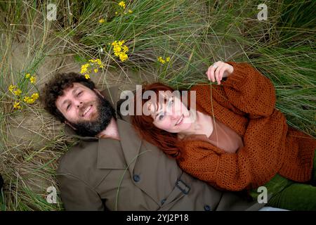 Lächelndes Paar, das auf dem Gras liegt, mit gelben Blumen um sie herum, in Herbstfarben gekleidet, Belgien Stockfoto