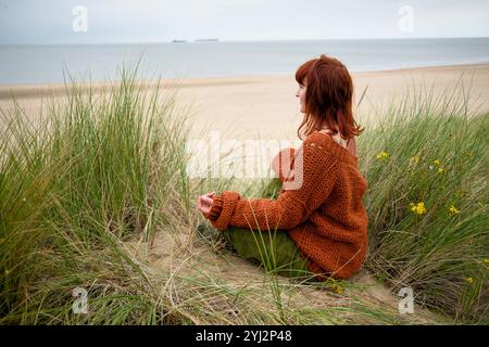 Frau, die in Sanddünen am Meer sitzt und an einem bewölkten Tag in den Horizont blickt, Belgien Stockfoto