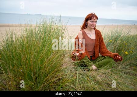 Frau, die Meditation in einer grasbewachsenen Düne übt, mit einem ruhigen Ausdruck und einem Schiff, das am Horizont sichtbar ist, Belgien Stockfoto