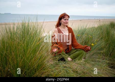 Frau, die in hohem Gras mit einer ruhigen Strandkulisse meditiert, Belgien Stockfoto