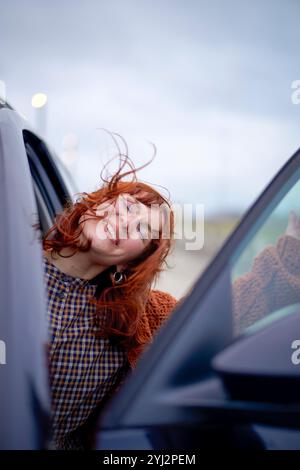 Rothaarige Frau lächelt mit geschlossenen Augen, lehnt sich aus einem Autofenster und weht im Wind die Haare. Stockfoto