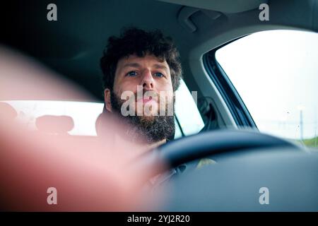 Bärtiger Mann mit lockigen Haaren, der ein Auto fährt, von der Beifahrerseite aus gesehen. Stockfoto