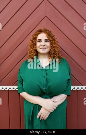 Portrait Plus Size schöne Frau mit lockigem rotem Haar im grünen Kleid, die in die Kamera vor buntem rotem Hintergrund der Wand schaut, Schönheit und Selbstvertrauen ou Stockfoto