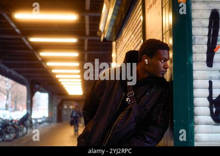 Ein junger Mann mit Ohrhörern lehnt sich an eine gekachelte Wand unter dem Schein von Deckenleuchten in einer U-Bahn-Station in Berlin Stockfoto