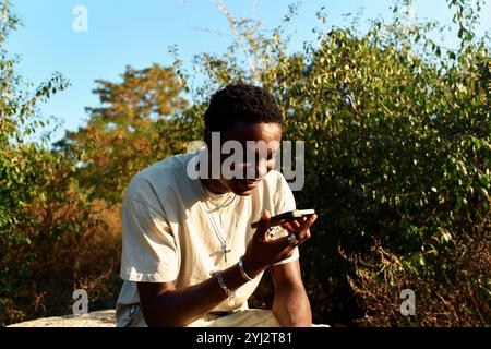 Ein Mann sitzt draußen auf einem Felsen und lächelt, während er mit seinem Smartphone spricht Stockfoto