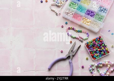 Bastelzubehör wie bunten Perlen, Buchstabenperlen, Schnur, Verschlüsse und Zangen sind auf einer rosafarbenen Fliesenoberfläche angeordnet, die für die Schmuckherstellung bereit ist Stockfoto