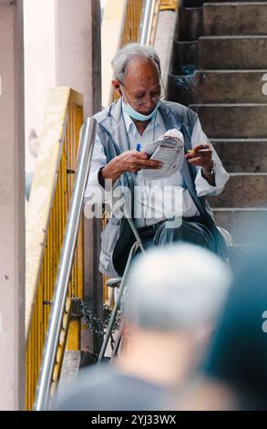 Ein älterer Mann sitzt auf einem Treppenhaus, in dem er eine Zeitung liest, während er eine Gesichtsmaske in der geschäftigen Umgebung Hongkongs trägt. Stockfoto