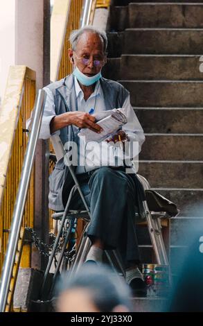 Ein älterer Mann sitzt auf einer Treppe, liest eine Zeitung, trägt eine Maske und Kopfhörer, in einer belebten Gegend von Hongkong. Stockfoto