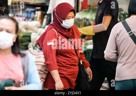 Eine Frau in rotem Hijab und Maske navigiert durch einen lebhaften Markt voller frischer Produkte und Menschen. Stockfoto