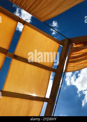 Sonnenlicht filtert durch ein texturiertes Stoffdach, von unten gesehen, eingerahmt von hölzernen Stützen vor einem leuchtend blauen Himmel und verstreuten weißen Wolken. Stockfoto