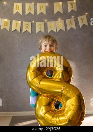 Glücklicher 8-jähriger Junge, der seinen Geburtstag feiert. Der Geburtstagsjunge steht im Zimmer und hält einen goldenen Festball in Form von Nummer acht in seinen Zähnen. Geburtsdatum Stockfoto