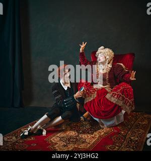 Ältere Frau und junger Mann vor dunklem Hintergrund. Die mittelalterliche Kaiserin sitzt auf einem Stuhl, während ein junger männlicher Diener ihren Musikspieler hält. Stockfoto