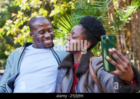 Ein lächelndes Paar nimmt Selfie in einem üppigen Stadtpark mit, eine fröhliche Beziehung im Freien Stockfoto
