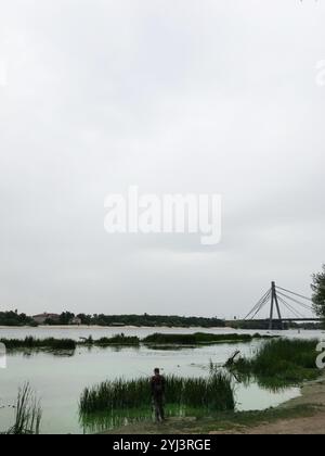 Ein einsamer Mann steht beim Angeln an einem ruhigen Flussufer, mit üppigem Grün und einer modernen Seilbrücke in der Ferne unter einem bewölkten Himmel. Stockfoto