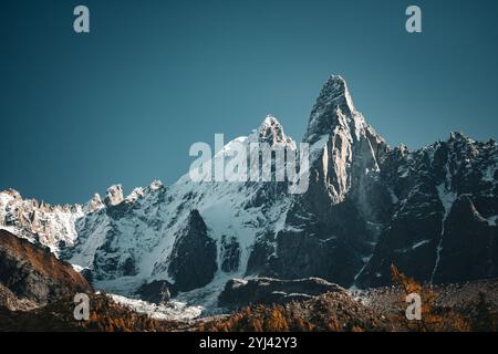 Schneebedeckte Berggipfel ragen über felsiges Gelände, beleuchtet von warmem Sonnenlicht, mit Herbstbäumen, die einen Kontrast unter einem klaren blauen Himmel bilden. Stockfoto