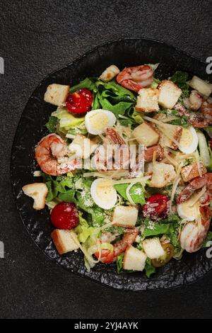 Caesar-Salat mit Tigergarnelen, Garnelensalat, Salatblättern, Wachteleiern, Kirschtomaten, parmesankäse, auf einem schwarzen Teller Stockfoto
