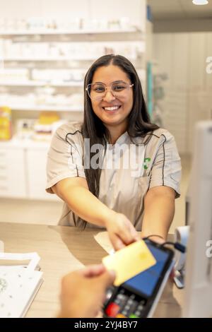 Vertikales Foto eines lächelnden Apothekers, der einen Kunden betreut, während er mit Kreditkarte am Tresen des Apothekengeschäfts bezahlt Stockfoto