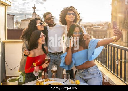 Glückliche, multiethnische Freunde, die während des Sonnenuntergangs auf einem Dach Selfie machen, mit Blick auf die Stadt Stockfoto