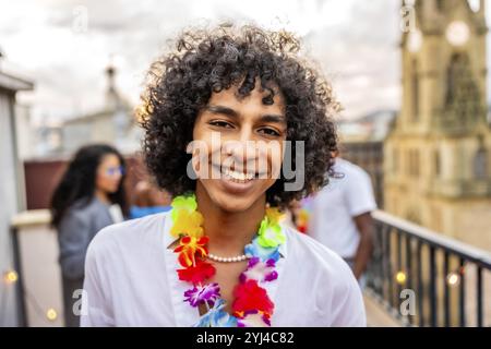 Glücklicher Mann mit lockigen Haaren und hawaiianischer Halskette, der beim Feiern auf dem Dach lächelt Stockfoto