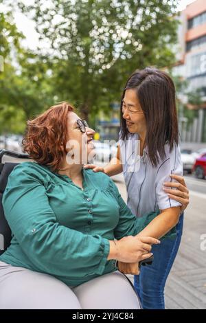 Vertikales Foto einer glücklichen Frau im Rollstuhl, die die Unterstützung einer multiethnischen Freundin erhält, die gemeinsam durch die Stadt spaziert Stockfoto