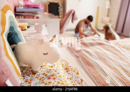 Ein weißes Einhorn Plüschtier sitzt auf einem Bett mit einem Regenbogenkissen im Schlafzimmer, verschwommene Figuren im Hintergrund Stockfoto