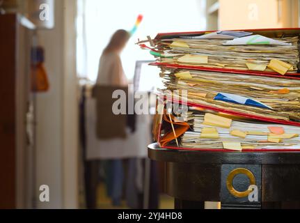 Haufen von Aktenmappen und verschwommenem Arbeitsplatz und unerkennbare Frau mit Reinigungsutensilien. Geschäftskonzept Doppelbelastung von Hausarbeit und Heim o Stockfoto