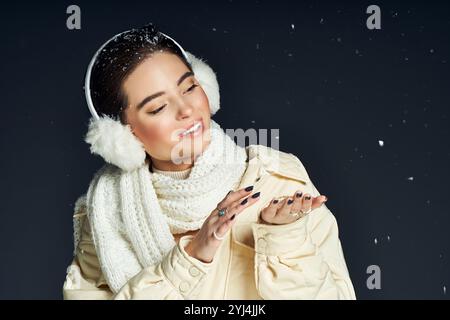 Eine lächelnde Frau spielt mit fallenden Schneeflocken, während sie warme Ohrenschützer und einen Schal trägt. Stockfoto