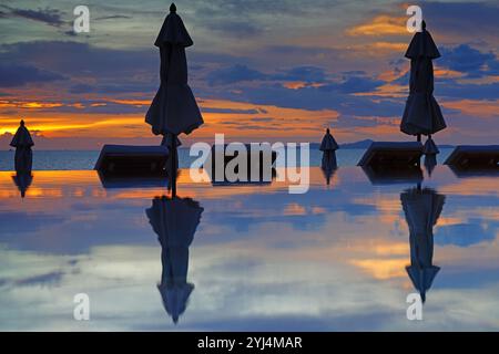 Geschlossene Sonnenschirme in einem Infinity-Pool mit Blick auf das Meer in einem dramatischen Sonnenuntergangshimmel Stockfoto