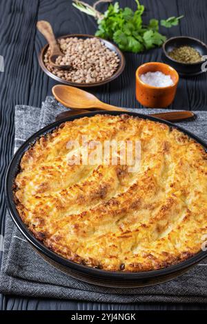 Veganer Hirtenkuchen in Backform auf schwarzem Holztisch mit Zutaten, Gewürzen, frischem Koriander und Holzlöffeln, vertikale Ansicht von oben, Nahaufnahme Stockfoto